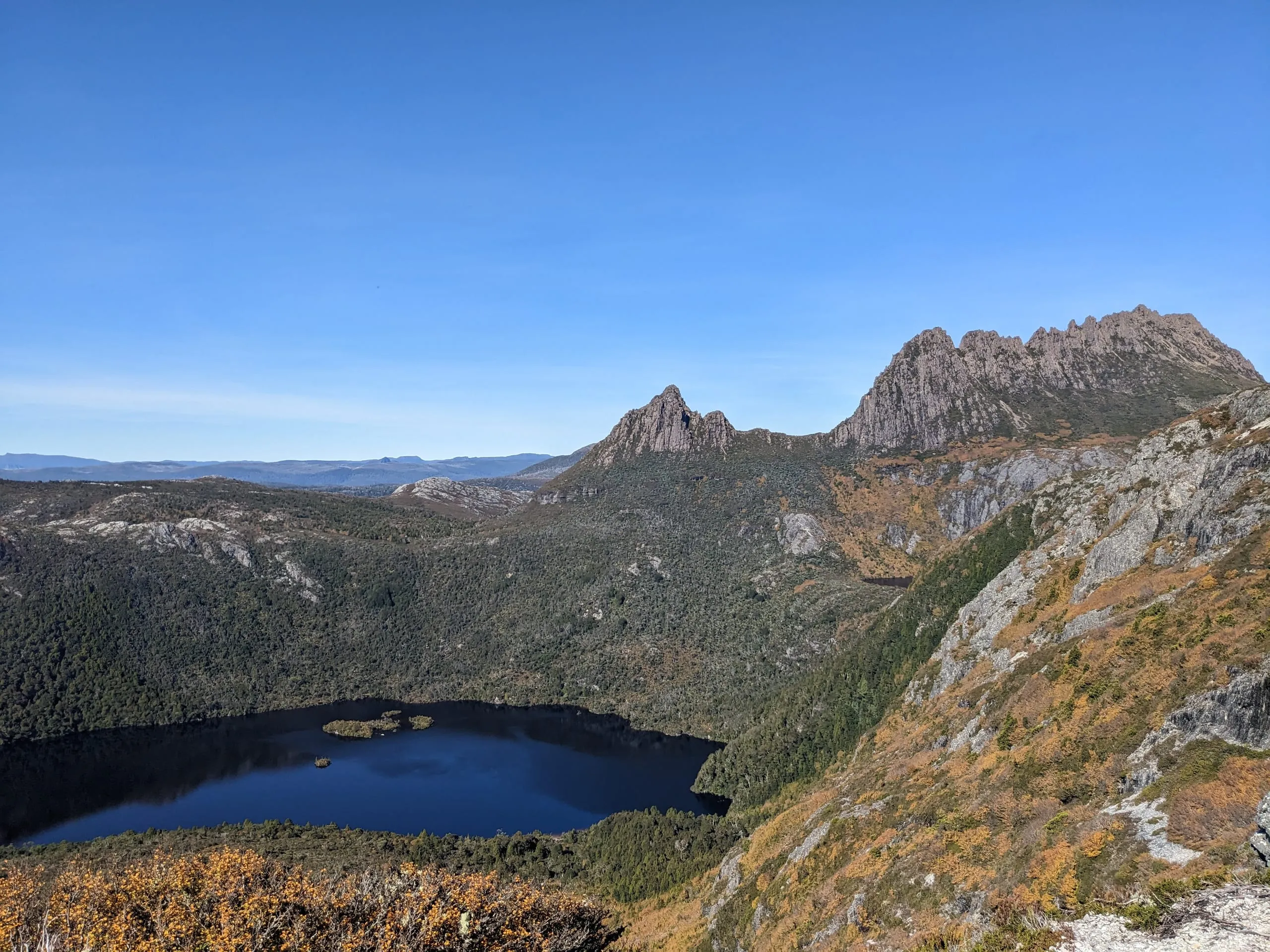 Cradle Mountain's jagged peaks above a dark lake ringed by autumn scrub, under a clear pale sky.