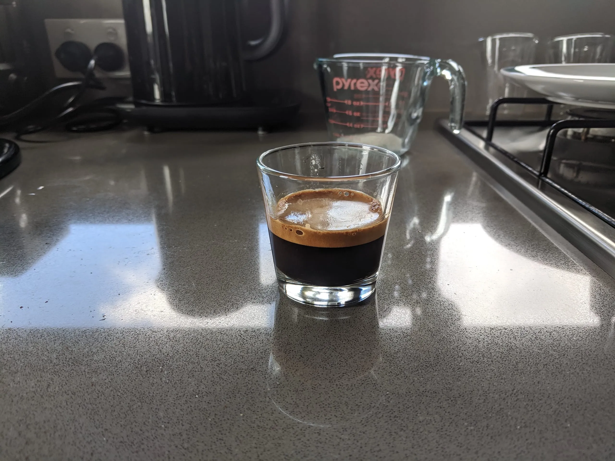 A glass of fresh espresso with thick crema on a kitchen bench, lit by a stripe of morning sun.