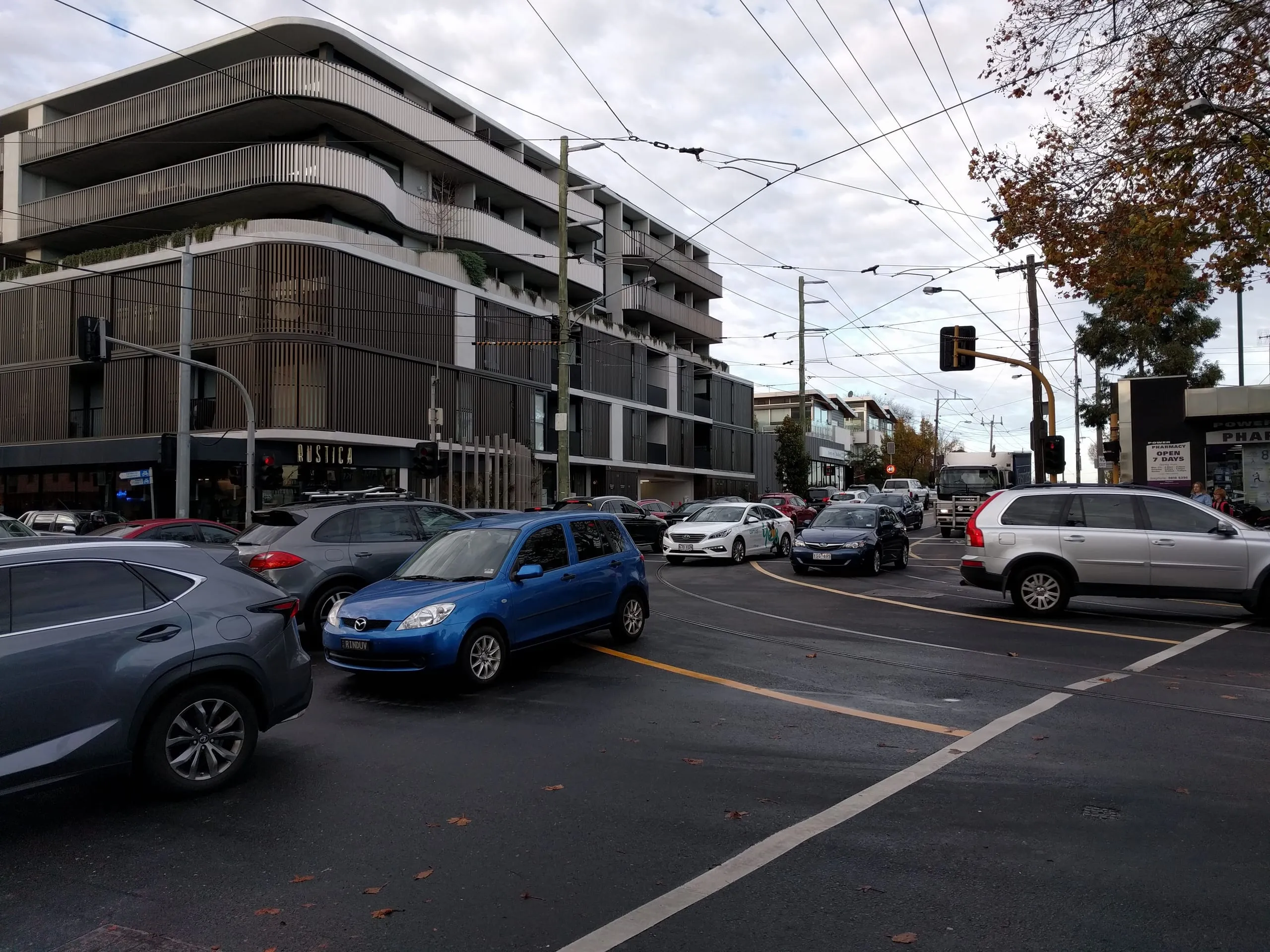 A grey city intersection under tram wires, cars queued in every direction, one bright blue hatchback mid-turn.