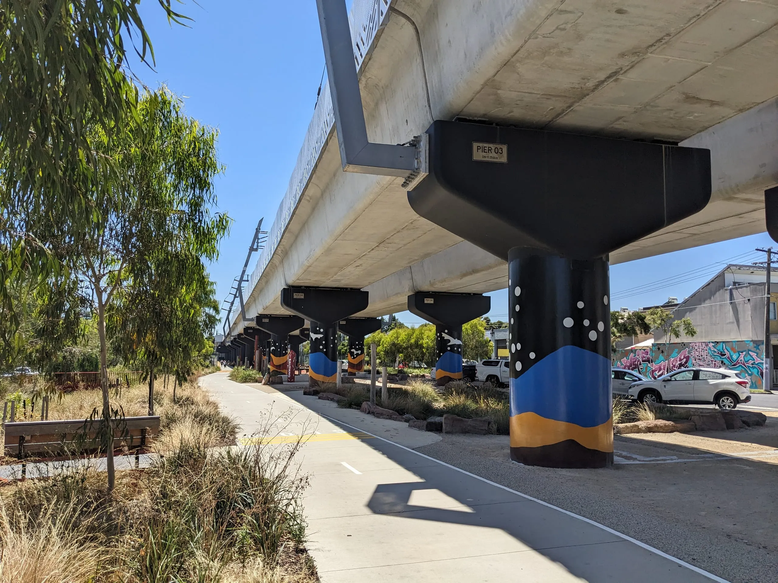 A walking path beneath an elevated rail line, its concrete pylons painted in colourful murals, gum trees alongside.
